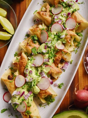 overhead of flautas on a large platter with avocado sauce on top